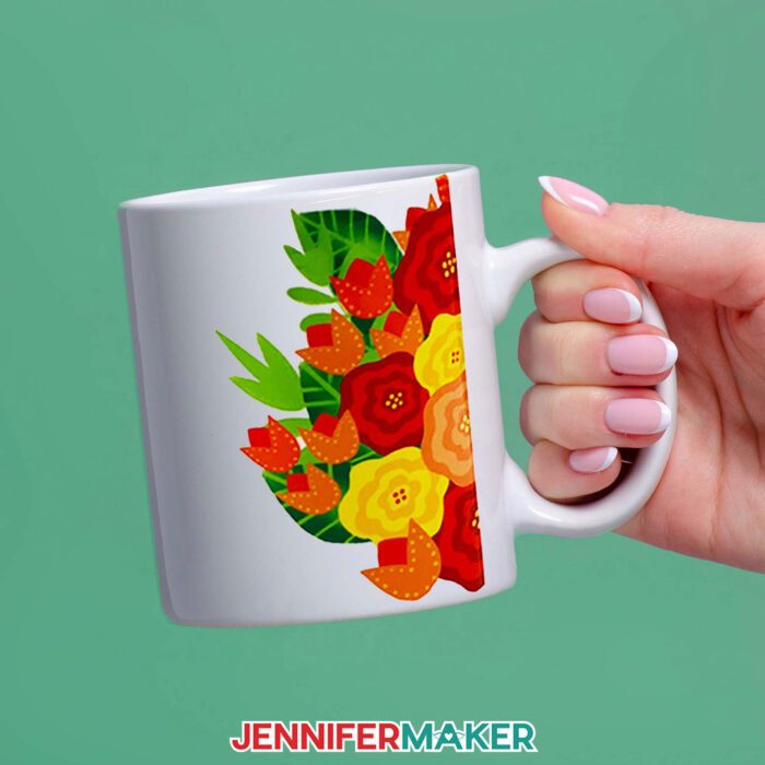 Sublimated floral mug, held against a background of green. The floral design goes around the handle, and is made up of red, orange, and yellow flowers. Learn how to sublimate mugs with JenniferMaker's tutorial!