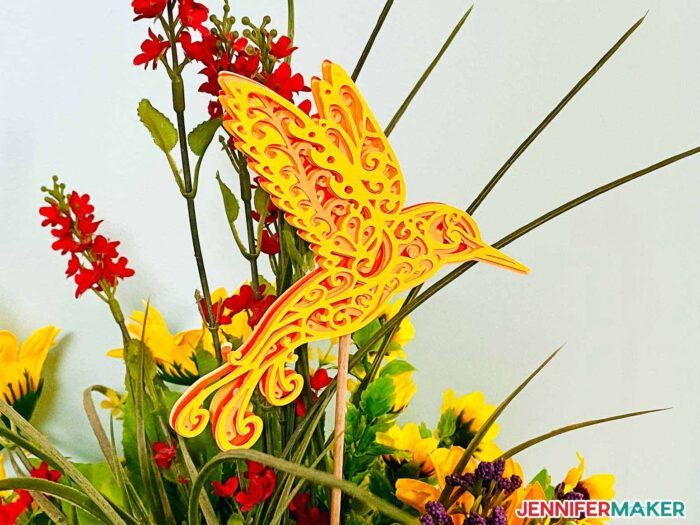 Layered cardstock hummingbird with intricate cuts on Cricut in a bouquet.