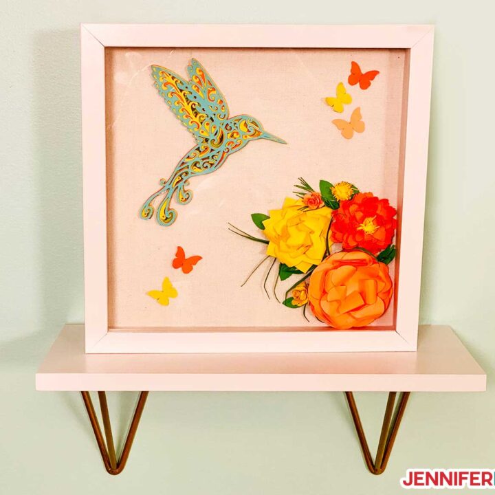 Shadow box with a layered cardstock hummingbird with intricate cuts on Cricut and paper decorations.