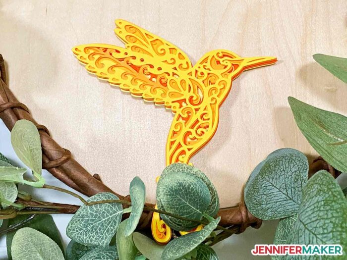 Layered cardstock hummingbird with intricate cuts on Cricut in in a wreath.