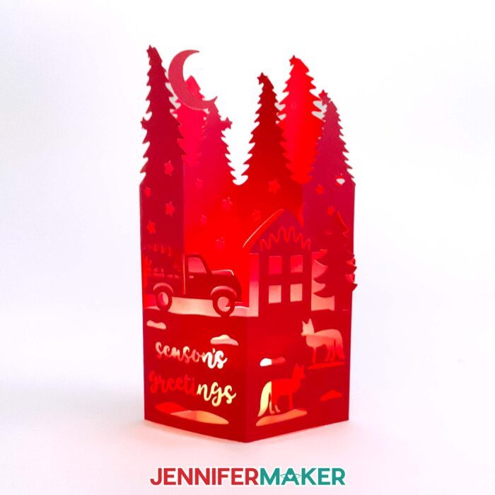 Make easy lantern pop-up cards with JenniferMaker's tutorial! A beautiful pop-up lantern cards with a moonlit night, a truck, a cottage, two foxes, and the message "Seasons Greetings" cut out, made from red cardstock, stands up, lit from the inside by a battery-operated tealight candle.