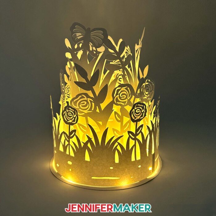 An illuminated white Layered Garden Lantern glows in a dark space, with intricate cutouts of roses, grass, butterflies, and dragonflies in a 3-tiered cylindrical shape