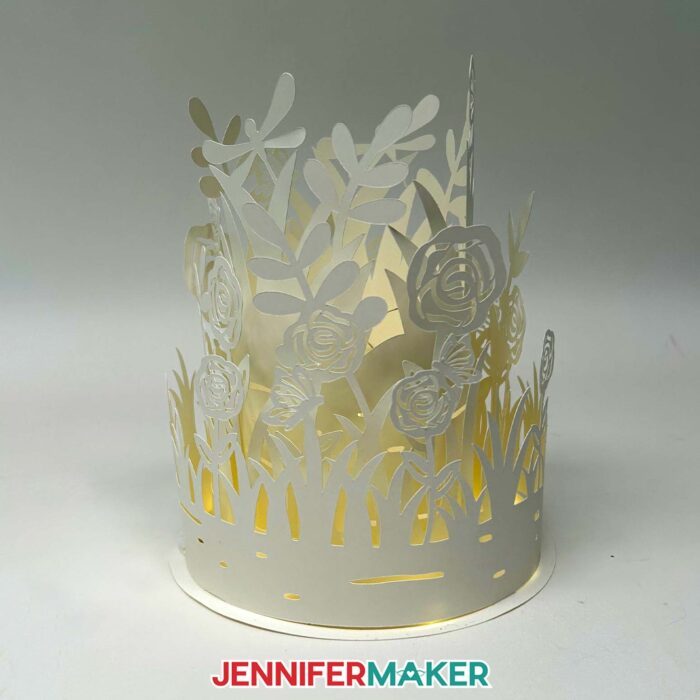 An illuminated white Layered Garden Lantern glows softly against a white backdrop, with intricately cut leaves, roses, grass, butterflies, and dragonflies in a 3-tiered cylindrical shape