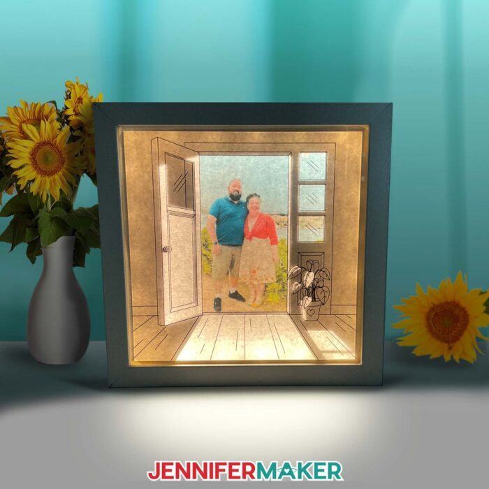 Make a light painting shadow box with JenniferMaker's tutorial! An illuminated shadowbox frame sits upon a table surrounded by sunflowers and a pretty blue backdrop. The shadowbox features a drawn doorway image with a real photo of Jennifer and Greg inside. The lights inside the box cast light rays across the floor of the image.