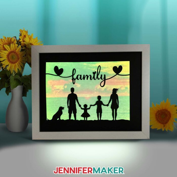 Make a light painting shadow box with JenniferMaker's tutorial! An illuminated shadowbox frame sits upon a table surrounded by sunflowers and a pretty blue backdrop. The shadowbox features a lit-up photo of a sunset, with silhouettes of a family of four (plus their dog) in the foreground. At the top is the word "family" in script, flanked by two hearts.