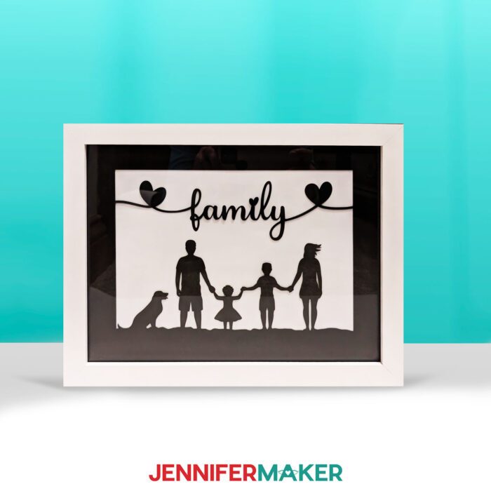 Make a light painting shadow box with JenniferMaker's tutorial! An illuminated shadowbox frame sits upon a table surrounded by sunflowers and a pretty blue backdrop. The shadowbox features silhouettes of a family of four (plus their dog) in the foreground. At the top is the word "family" in script, flanked by two hearts. The sunset photo is hidden because the light in the shadowbox is off.