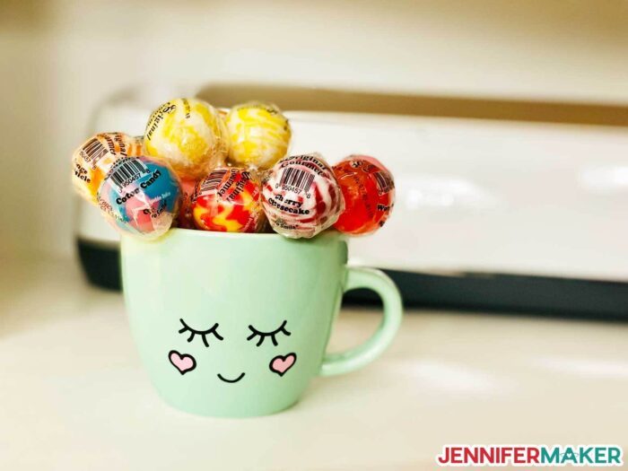 Several multicolored round lollipops in a light green mug with a face decoration on a table in front of a Cricut.