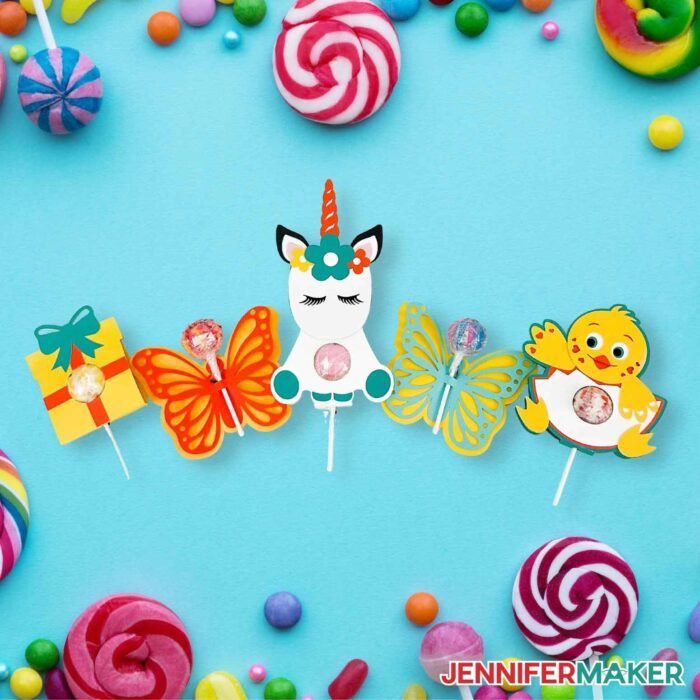Present, unicorn, chick, and butterfly cardstock DIY lollipop holders on a blue background surrounded by candy.