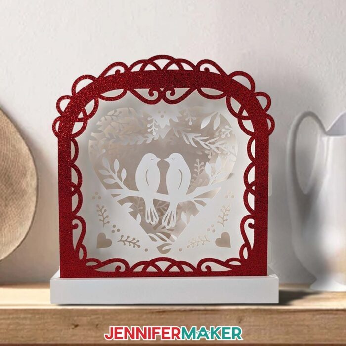 Make a love paper lantern with Jennifer Maker! A white cardstock lantern with love birds in the center and a red glitter frame sits on a mantel.
