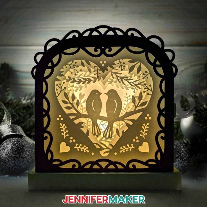 Learn how to make a Love Paper Lantern with JenniferMaker's tutorial! An intricately layered paper lantern featuring two birds, hearts, leaves, and black details sits illuminated in a wintery space.