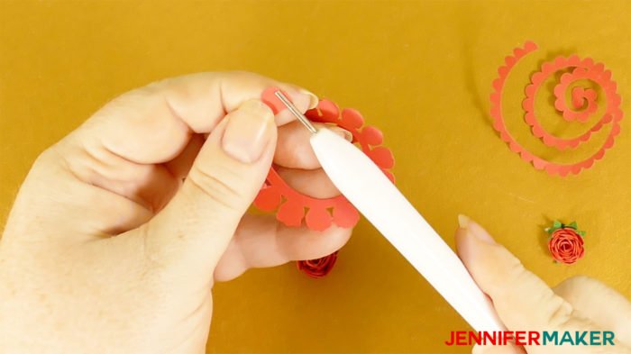 The tip of a red spiral petal pattern is inserted into the tip of a quilling tool to make miniature paper roses