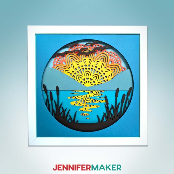 Get Jennifer Maker's best paper mandala tips! A cardstock mandala featuring a sunset over a lake hangs on a blue backdrop.