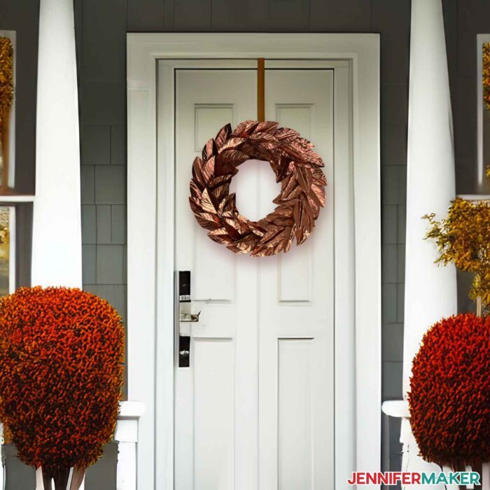 Coppery metal wreath with leaves cut on a Cricut hung on an emerald white front door with fall decor surrounding it.