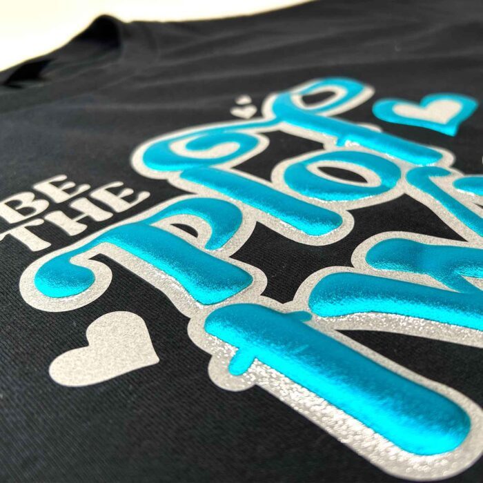Extreme close-up of silver glitter vinyl layered under turquoise puff vinyl decorate a black shirt, with the quote "Be The Plot Twist" from the Puff Vinyl Tutorial