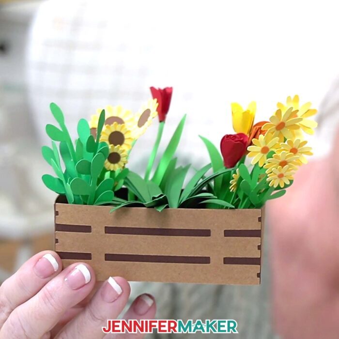 A hand holds a mini paper crate full of mini flower arrangements from JenniferMaker's Mini Paper Flower Tutorial