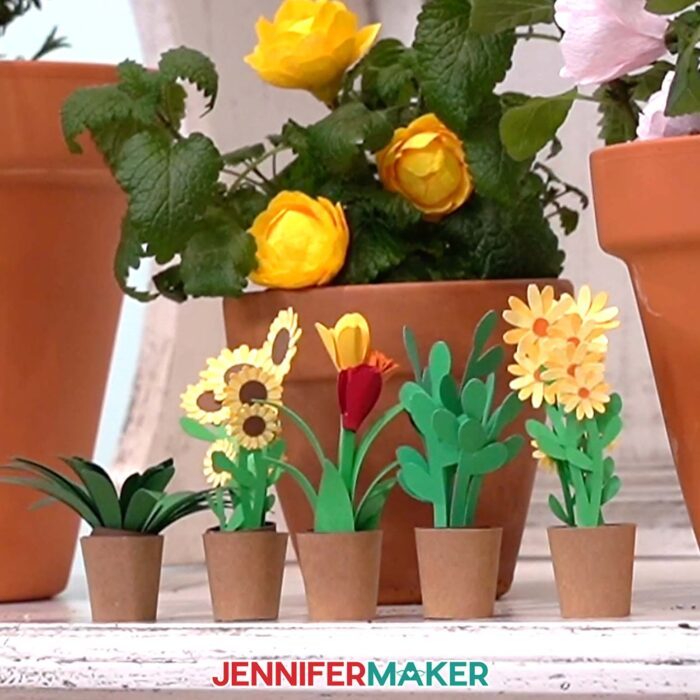 Collection of mini paper flowers and flower arrangements on display in paper pots in front of a large, real pot of flowers, all made from JenniferMaker's Mini Paper Flower Tutorial