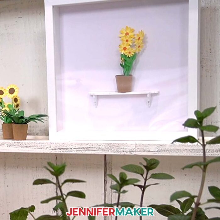 A shadow box sits on display, with a mini potted sunflower flower arrangement sitting on a miniature shelf from JenniferMaker's Mini Paper Flower Tutorial