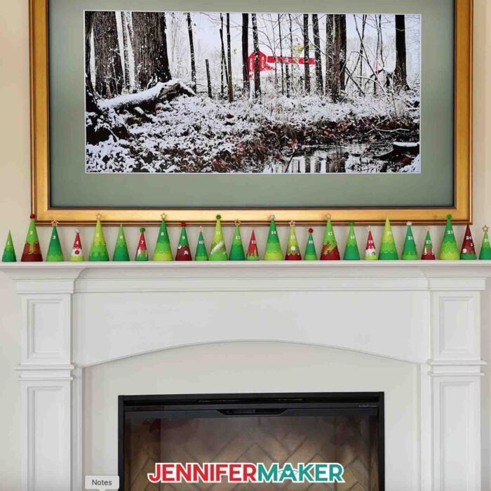 A full 25-tree Minimalist Tree Advent Calendar project on a fireplace mantel, made up of colorful cone-shaped tree boxes made from cardstock, under a large framed photo of a snowy Maker Farm through trees