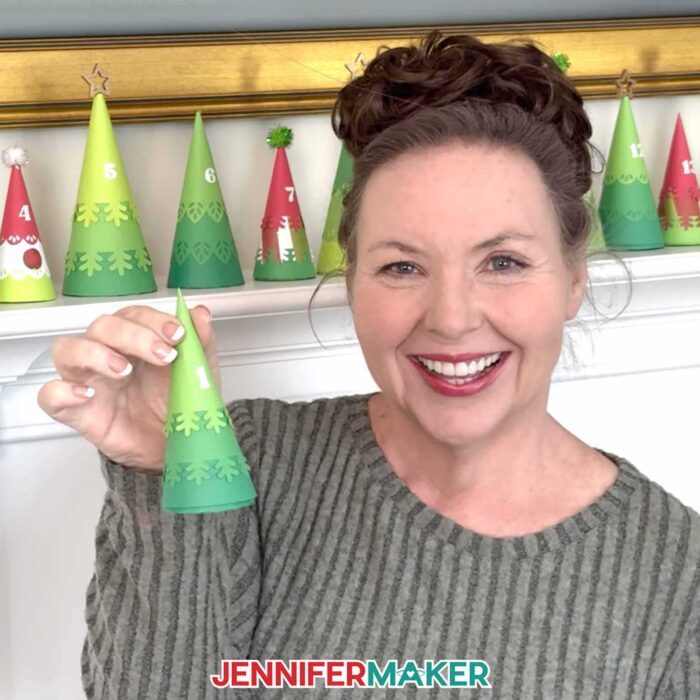 Jennifer Maker shows off a small green Minimalist Tree Advent Calendar project box in front of her fireplace mantel, which made up of colorful cone-shaped tree boxes made from cardstock