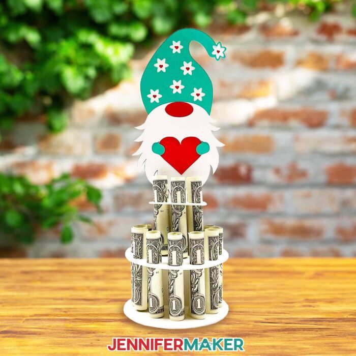 Colorful money cake decoration holding rolled dollar bills in a column, topped with a cute gnome with a teal hat holding a red heart, made using a JenniferMaker SVG file.