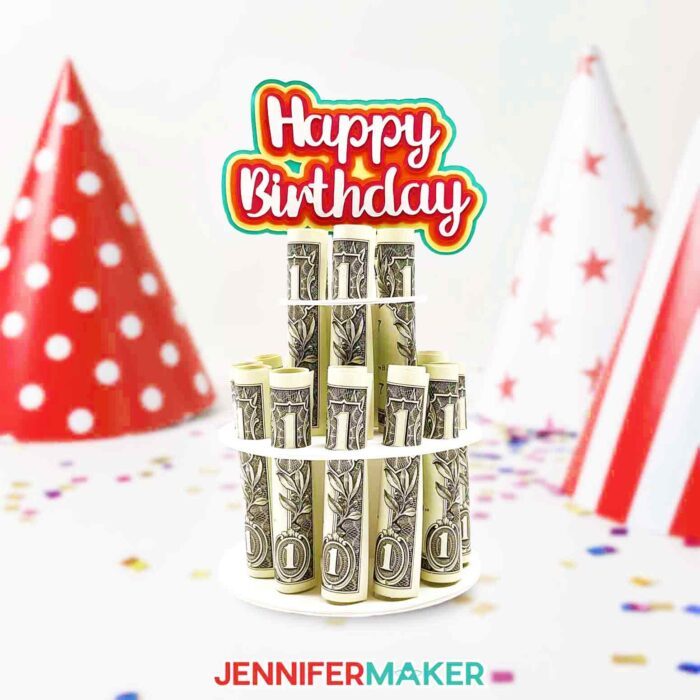 Colorful two-tier money cake decoration holding rolled dollar bills in a column, topped with a multicolor Happy Birthday sign, made using a JenniferMaker SVG file and tutorial.