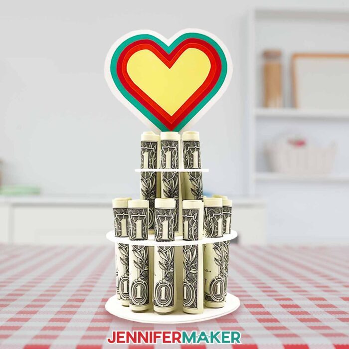 Colorful money cake decoration holding rolled dollar bills a in two-tier column, topped with a multicolor heart, made using a JenniferMaker SVG file.