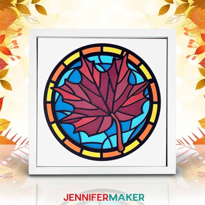 A shadow box frame holding layered paper in a stained glass inspired design featuring a red maple leave on a blue blackground.