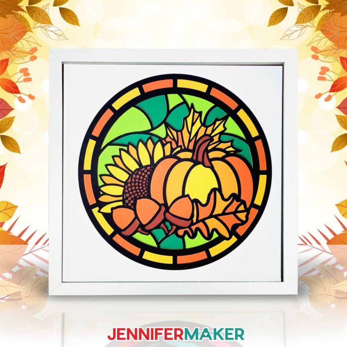A shadow box frame holding layered paper in a stained glass inspired design featuring a sunflower, leaves, a pumpkin, and acorns.