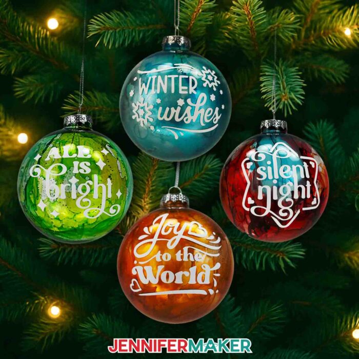 Four alcohol ink ornaments with white vinyl messages hanging on a tree: "Winter Wishes" on a blue ornament, "All Is Bright" on a green ornament, "Silent Night" on a red ornament, and "Joy to the World" on an orange ornament.