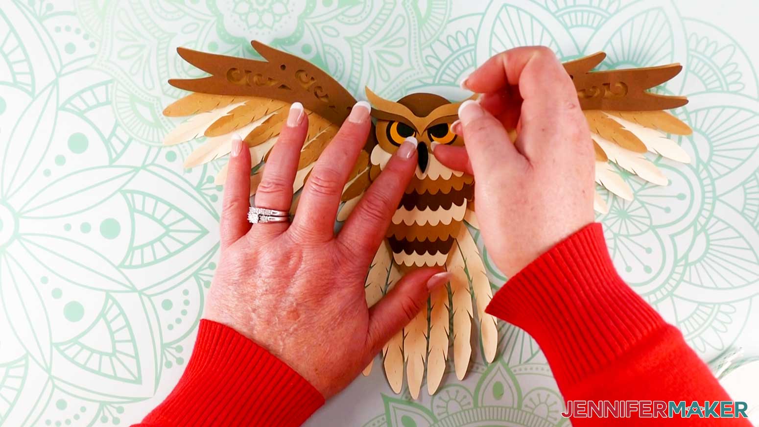 An overhead photo showing two hands above the assembled layered owl aligning and placing the small beak piece on top of the center of the face piece