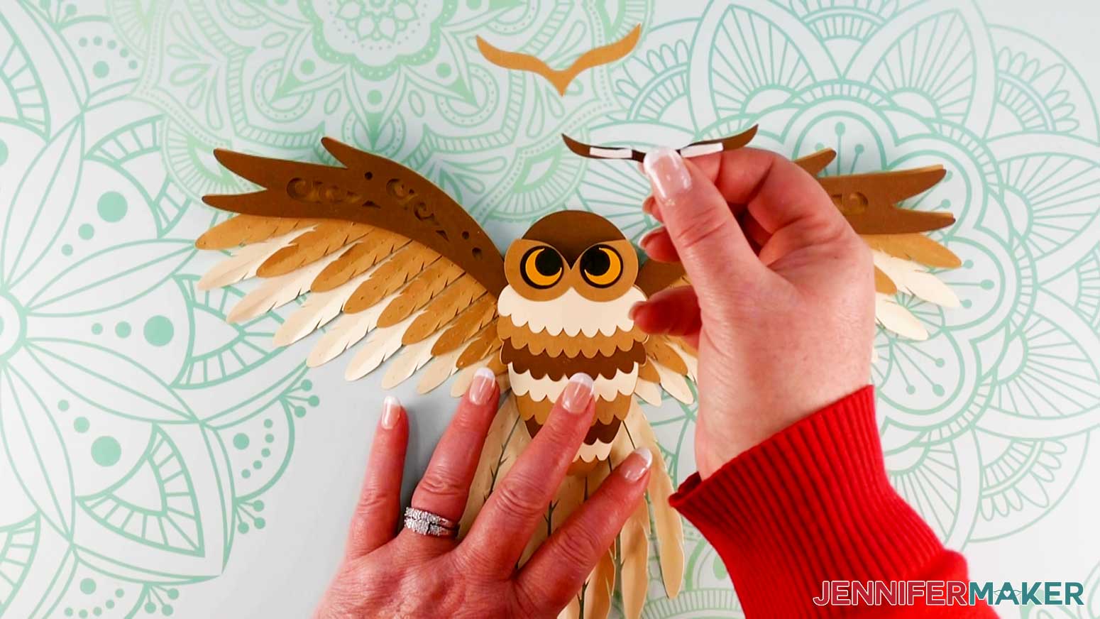 An overhead photo showing one hand holding the dark brown tuft piece with two thin adhesive foam strips attached to the back, while another hand holds the assembled layered owl in place on the work surface