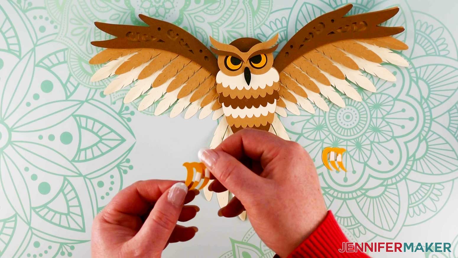 An overhead photo showing two hands holding the left talon piece with two small strips of adhesive foam tape applied to the back of it and the assembled layered owl and right talon piece laying on the work surface below