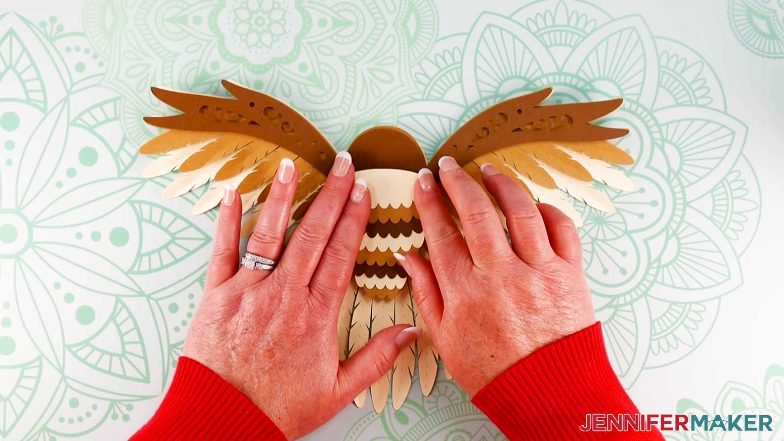 An overhead photo showing two hands aligning the topmost cardstock piece of the layered owl body feathers to the other seven body feather pieces, all of which are glued to the center of the large wing pieces of the design