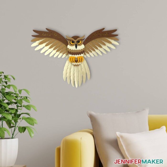 Papercraft layered owl SVG made of tan, brown, and yellow cardstock with spread wings mounted on a gray wall by a yellow couch.