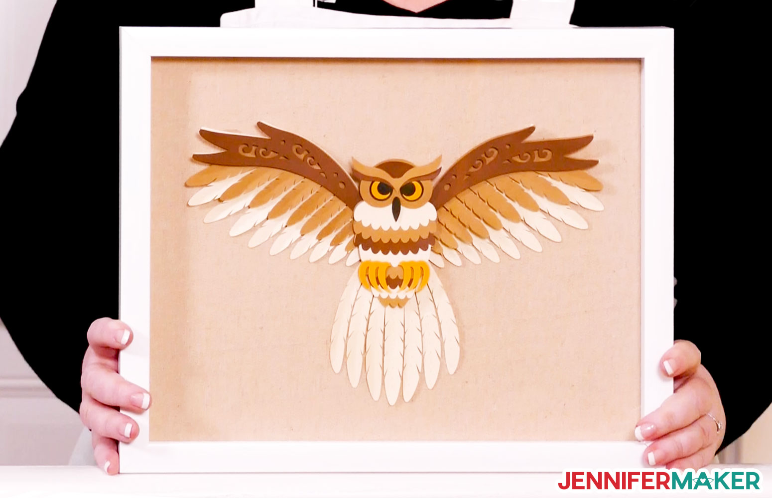 Papercraft layered owl svg made of tan, brown, black, and yellow cardstock with spread wings in a shadow box being held by Jennifer Maker