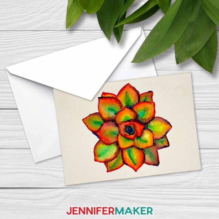 A notecard with a watercolored painted succulent rests on top of an envelope, on a whitewashed wooden surface with plants nearby. Learn how to paint watercolor plants with a Cricut! With JenniferMaker's tutorial, you can learn how to paint with watercolor and Cricut!