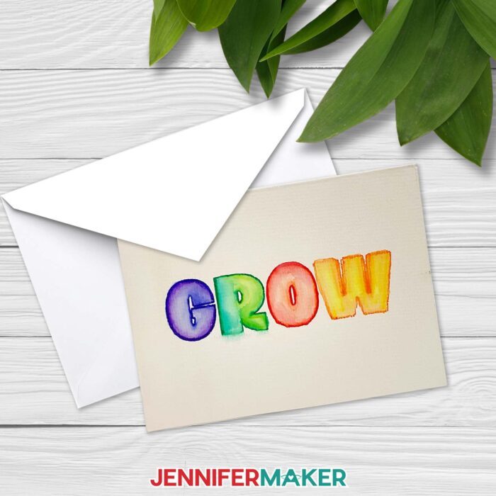 A notecard with a watercolored painted word "GROW" rests on top of an envelope, on a whitewashed wooden surface with plants nearby. Learn how to paint watercolor plants with a Cricut! With JenniferMaker's tutorial, you can learn how to paint with watercolor and Cricut!