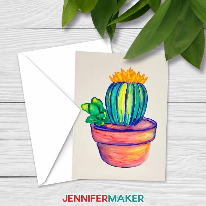 A notecard with a watercolored painted potted cactus rests on top of an envelope, on a whitewashed wooden surface with plants nearby. Learn how to paint watercolor plants with a Cricut! With JenniferMaker's tutorial, you can learn how to paint with watercolor and Cricut!