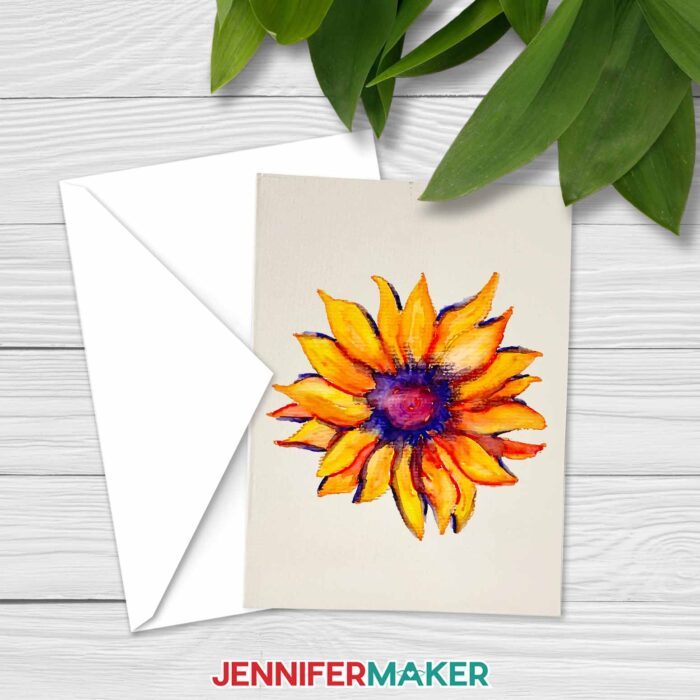 A notecard with a watercolored painted sunflower rests on top of an envelope, on a whitewashed wooden surface with plants nearby. Learn how to paint watercolor plants with a Cricut! With JenniferMaker's tutorial, you can learn how to paint with watercolor and Cricut!