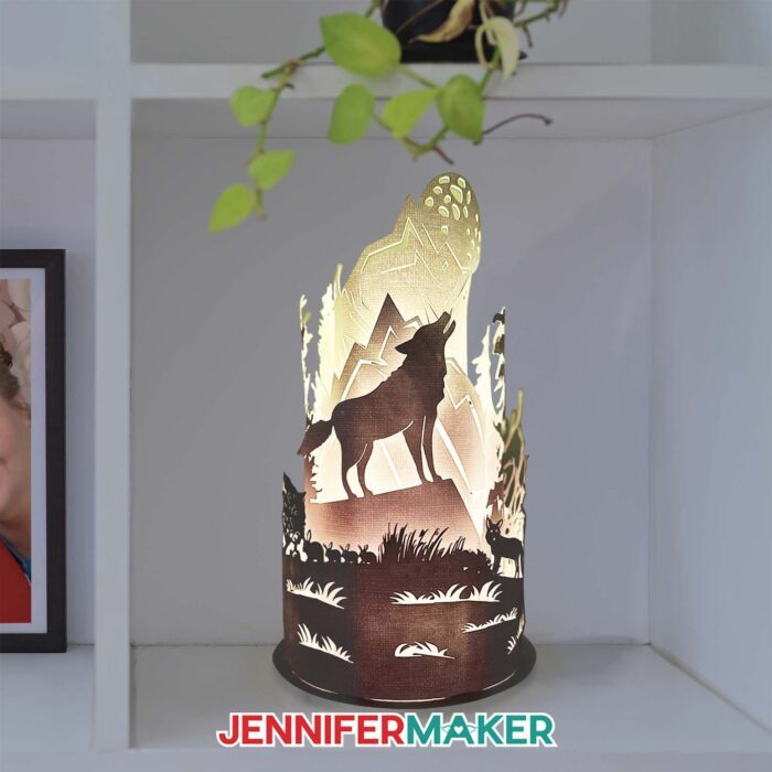 A rounded paper cut lamp displaying a wolf howling at the moon surrounded by mountains, trees, and wildlife, lit from within on a white shelf.
