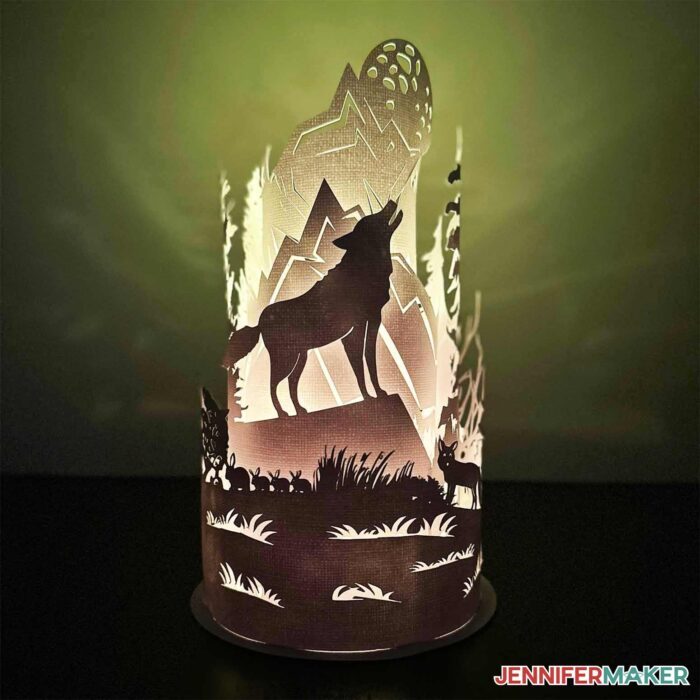 A rounded paper cut lamp displaying a wolf howling at the moon surrounded by mountains, trees, and wildlife, lit from within on a green background.