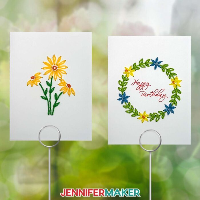 Make Paper Embroidery Cards! Two embroidered greeting cards, one with yellow daisies and one with a floral wreath with "Happy Birthday" in script inside it are on display on silver holders outdoors