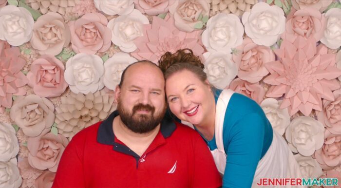 Two people posing happily in front of a paper flower backdrop featuring pink and cream handmade flowers.