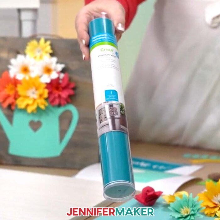 Learn how to create a beautiful paper flower sign with JenniferMaker's tutorial! Jennifer holds a teal colored tube of permanent vinyl over her work surface. 
