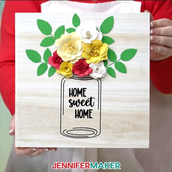 Learn how to create a beautiful paper flower sign with JenniferMaker's tutorial! A vinyl mason jar design reading "Home Sweet Home" decorates a whitewashed wooden panel with yellow and red flowers with green leaves. 
