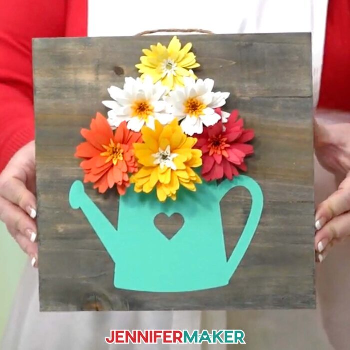 Learn how to create a beautiful paper flower sign with JenniferMaker's tutorial! A vinyl watering can design with a heart decorates a dark wooden panel with yellow, white, and red flowers. 