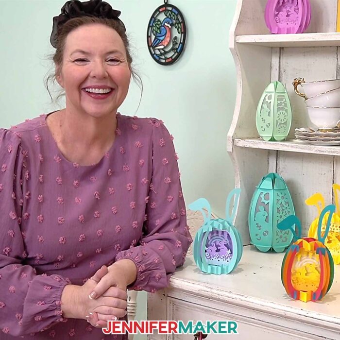 Jennifer Maker stands in front of a shelf filled with Paper Pop Up Easter Egg projects, smiling 