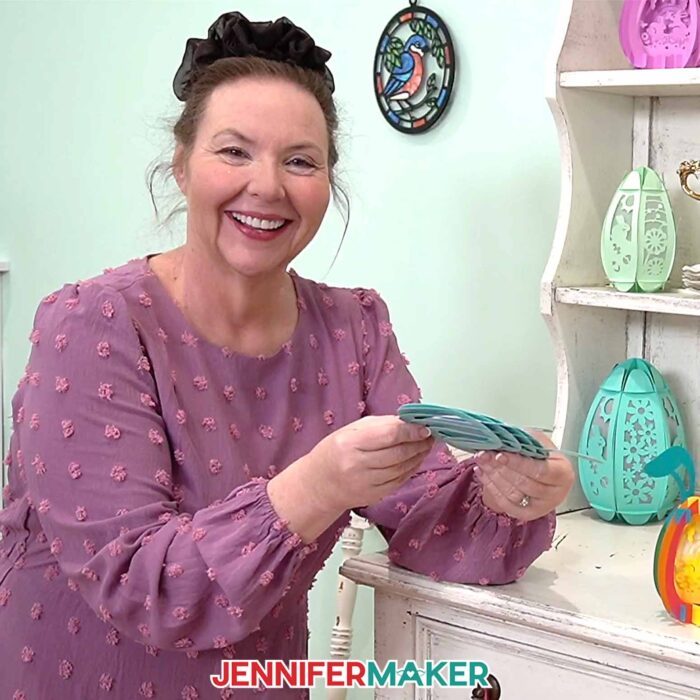 Jennifer Maker stands in front of a shelf filled with Paper Pop Up Easter Egg projects, demonstrating how to fold the project flat for mailing or storage