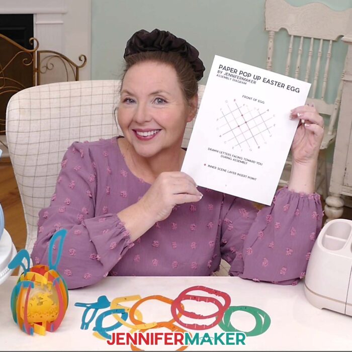 Jennifer Maker sits at her craft table, showing off a Paper Pop Up Easter Egg assembly diagram sheet, with pieces of a pop up egg on the table in front of her