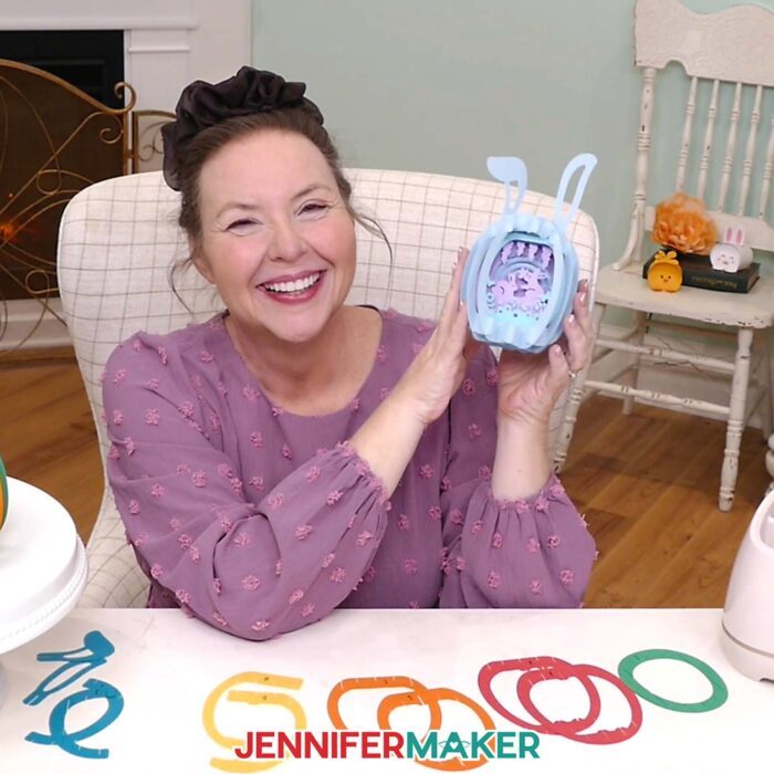 Jennifer Maker sits at her craft table, showing off a Paper Pop Up Easter Egg project, with pieces of another pop up egg on the table in front of her